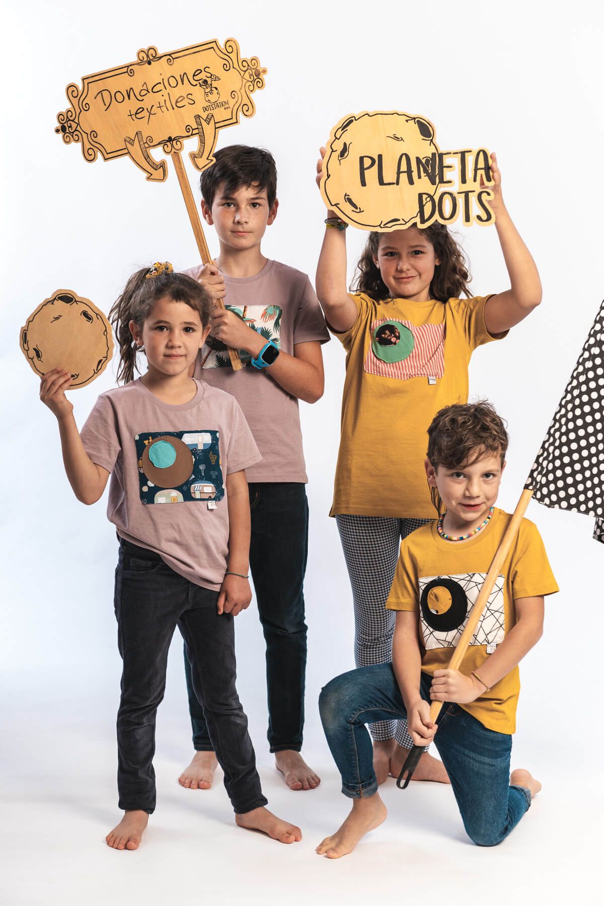 camiseta Kriptón Infantil (1) Niños posando con ropa de Planeta Dots, banderines de lunares y sujetando carteles que dicen "Donaciones textiles" y "Planeta Dots"
